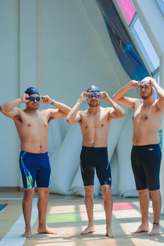 Three shirtless men with swim caps and goggles prepare by the indoor pool. Ready for a swim match.